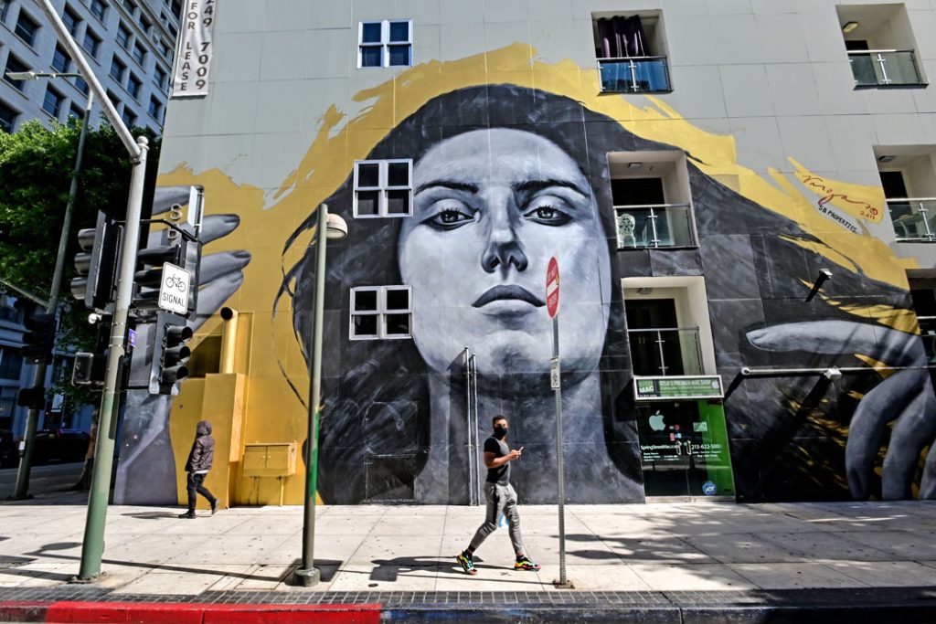 A man with mask walks past a mural of a woman on an empty street due to the COVID-19 outbreak in Los Angeles, March 2020/ Getty Image