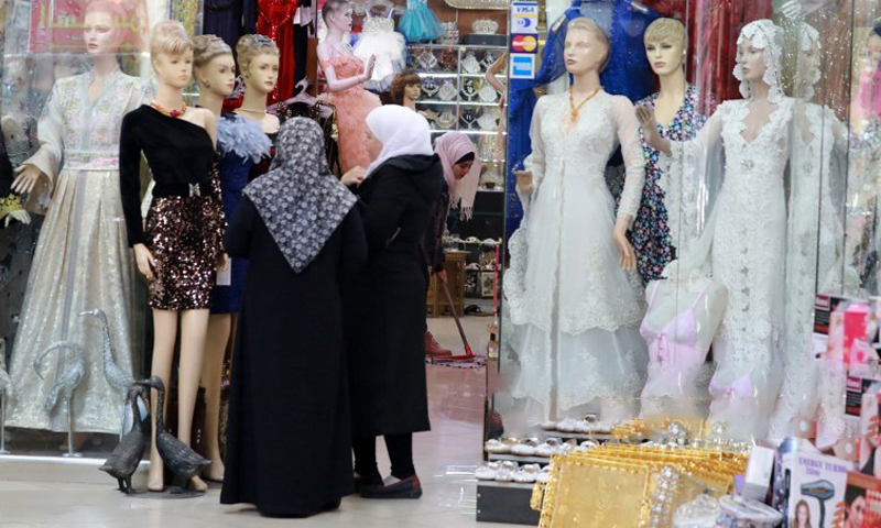 نساء سوريات ينظرن إلى فساتين معروضة أمام متجر لبيع الملابس في سوق الحميدية في المدينة القديمة بالعاصمة السورية دمشق - 3 كانون الأول 2019 (AFP)